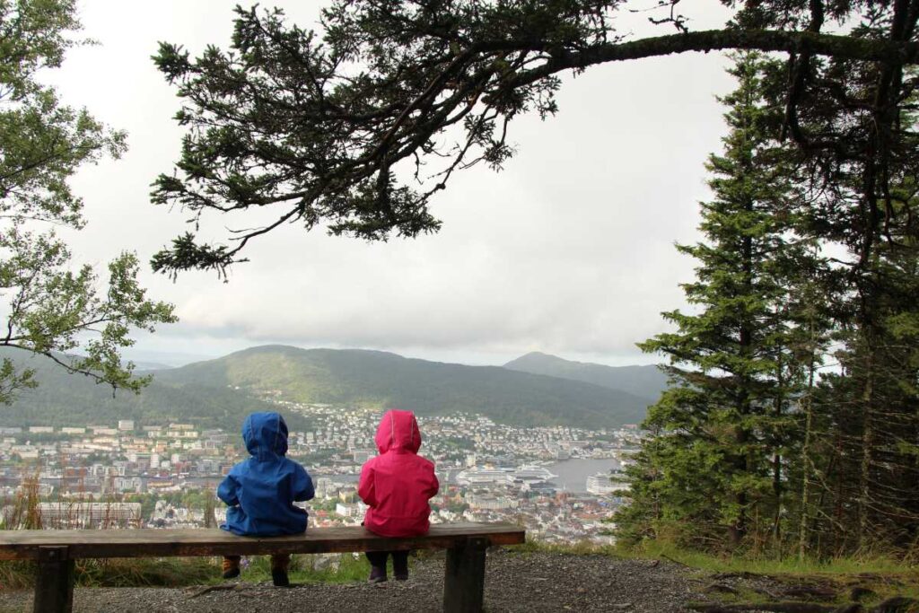 Two young children in rain gear sit on a bench overlooking Berrgen, Norway in summer.