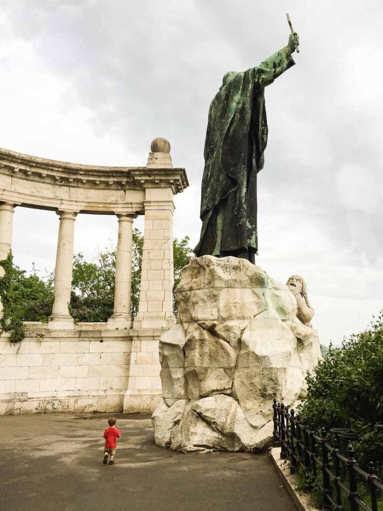 one of the Brewer kids, from the Family Can Travel blog, stands below the St. Gerard of Csanád Monument in Budapest, Hungary.