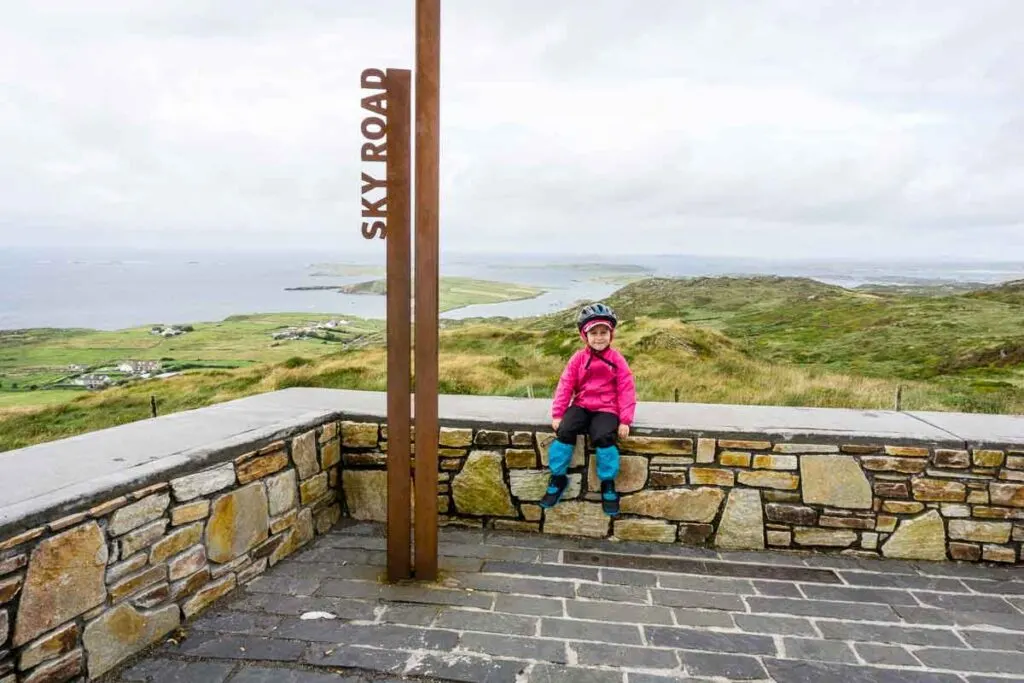 image of girl sitting on rock walk with ocean in background and next to sign that says Sky Road