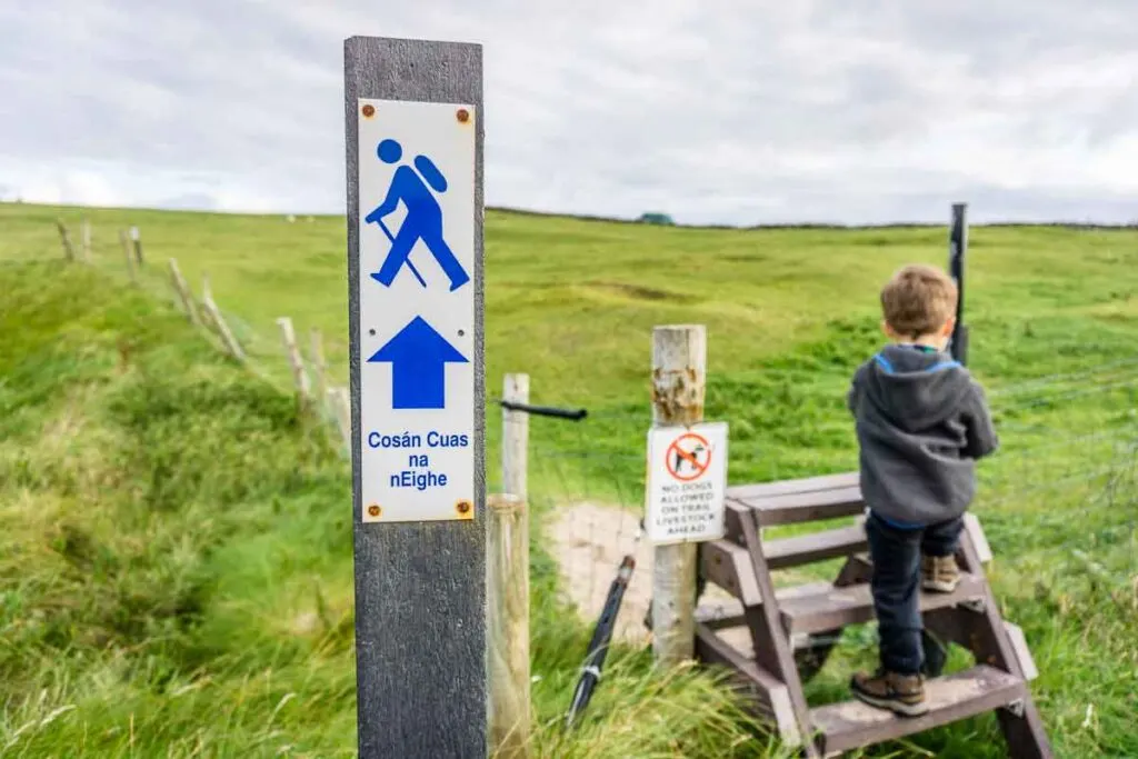 Image of Clogher Beach loop trail marker with boy in background climbing up ladder over a fence on Dingle Peninsula