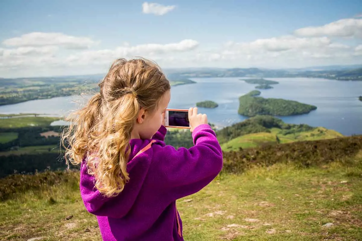 image of girl taking picture with a phone at the top of conic hill which is one of the best Loch Lomond walks to do with kids