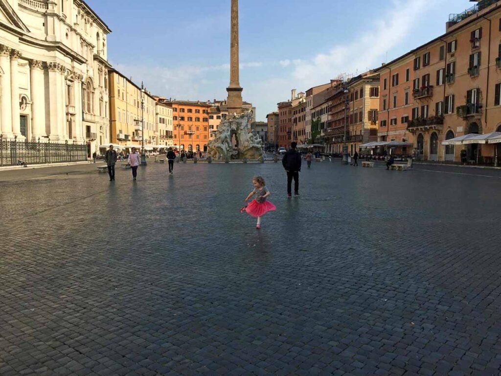A 3-year old girl in a bright pink skirt dances in an empty plaza during a family trip to Rome.