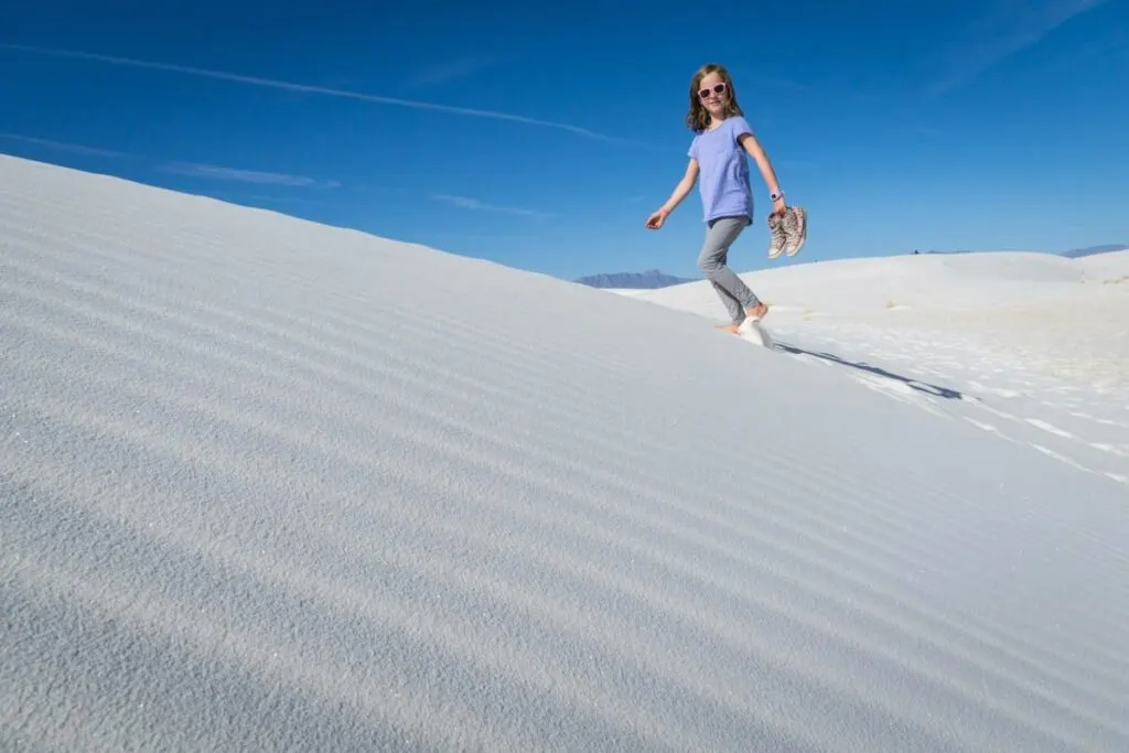 a young hiker carries her high-tops as she walks barefoot through the sand on the Alkali Trail