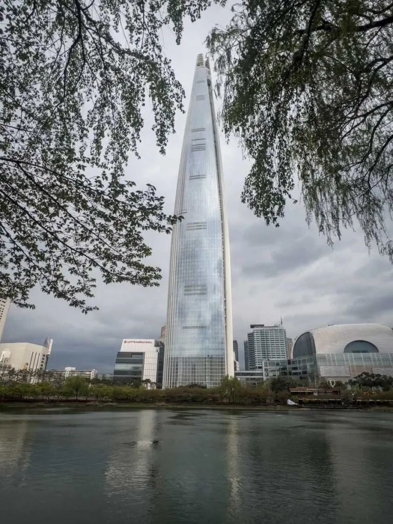 The Lotte World Tower is framed in-between two trees behind a nearby lake.