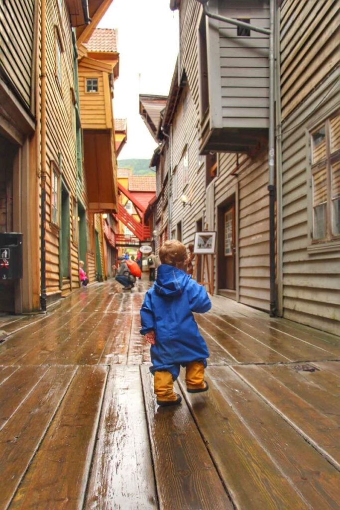 a 2-year old boy in a rain suit walks down a wet alleyway while on a family trip to Bergen, Norway.
