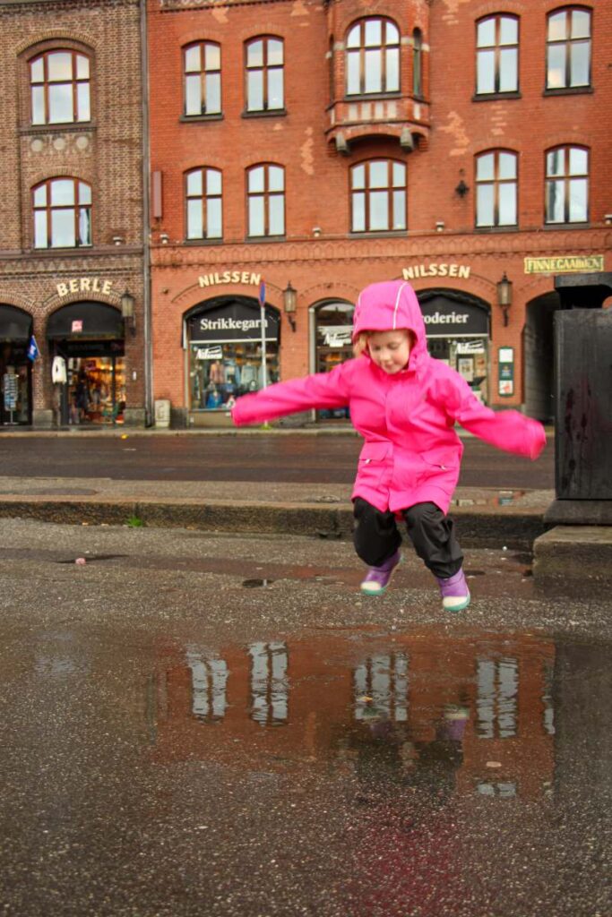 a 4-year old girl in a pink rain jacket jumps in a big rain puddle near Bryggen while on a family trip to Bergen.