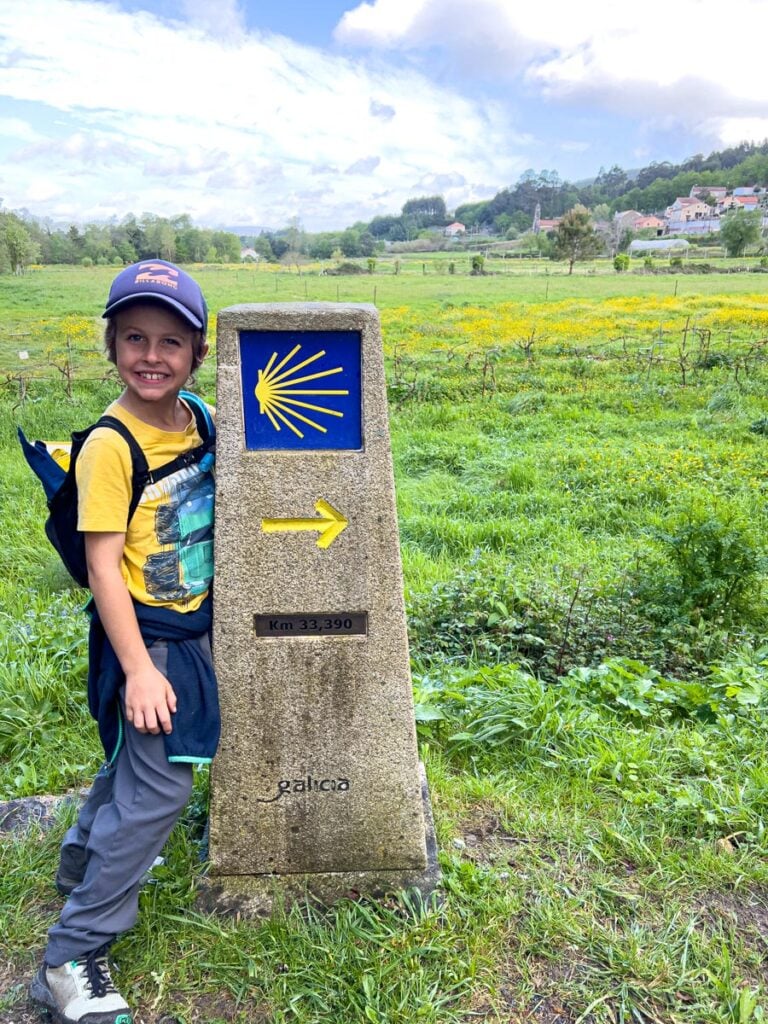 An 8-year old boy hiking the Portuguese Coastal Camino Route with his family poses next to a trail marker which shows it is only 33 km left to Santiago de Compostela.