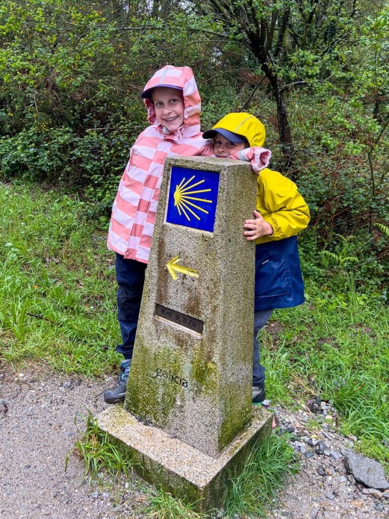 Two kids on a family pilgrimage to Santiago de Compostela pose next to the trail marker which shows only 10 km left in their journey.