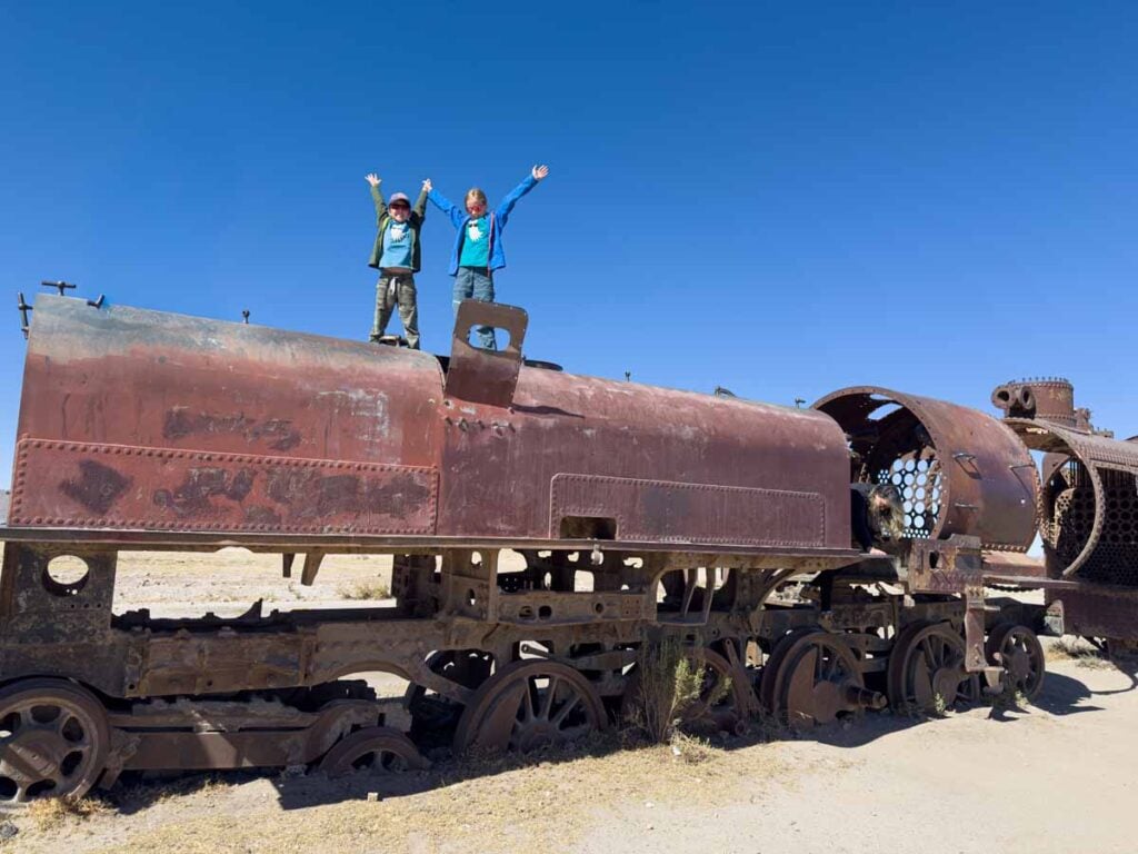 two kids stand atop a rusted out train engine at the famous train graveyard in the Uyuni Salt Flats in Bolivia.