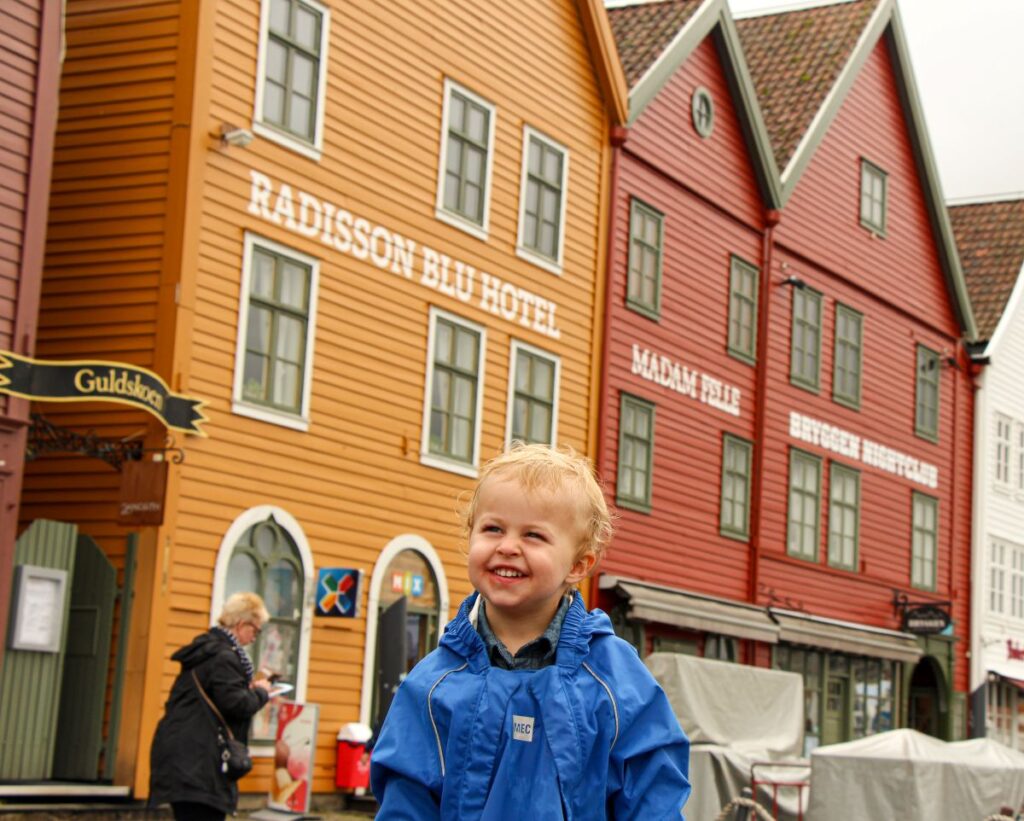 a toddler in rain gear is all smiles while visiting Bryggen on a family trip to Bergen, Norway.