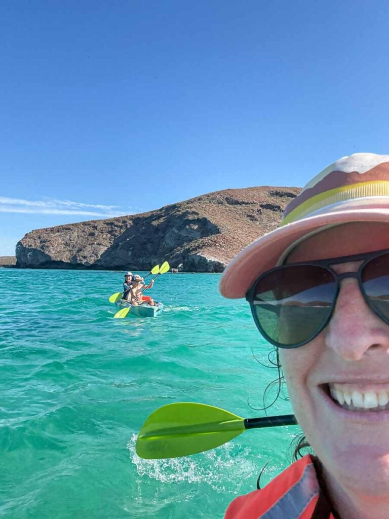 a selfie of Celine Brewer, from FamilyCanTravel.com, with her kids in the background on a family kayaking tour at Playa Balandra.