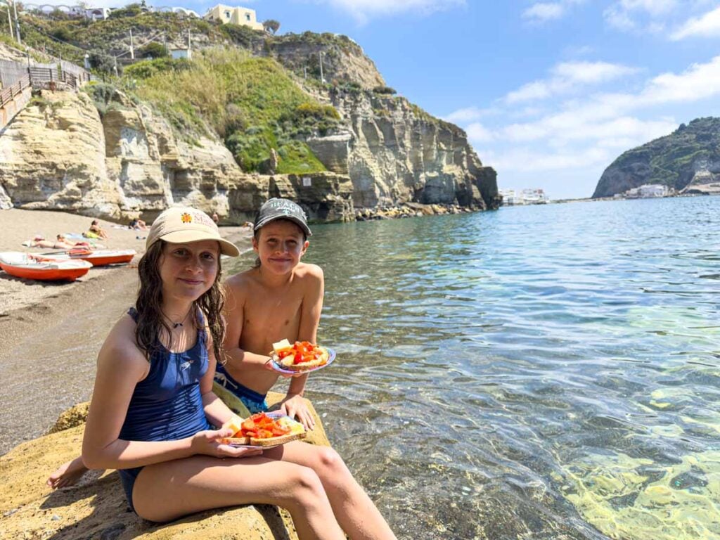 The Brewer kids, from the Family Can Travel blog, eat homemade Bruschetta on the beach in Ischia, Italy after a kayaking tour.