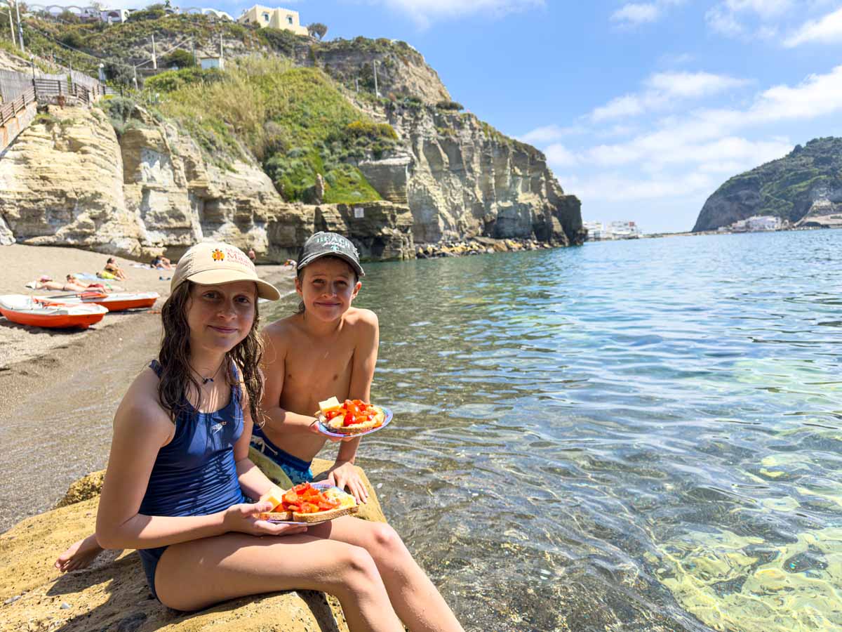 The Brewer kids, from the Family Can Travel blog, eat homemade Bruschetta on the beach in Ischia, Italy after a paddleboarding tour.
